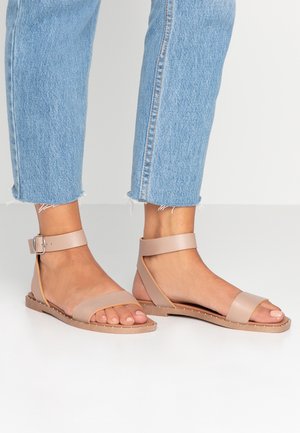 Pieds portant des sandales beige clair à bout ouvert avec bride à la cheville, assorties à un jean denim bleu clair coupé, sur une surface blanche.
