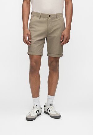 Bărbat purtând pantaloni scurți bej suflecați, șosete albe până la gleznă, adidași albi cu dungi negre, stând pe un fundal simplu și deschis la culoare.