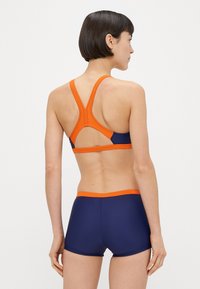 Μπλε ναυτικό αθλητικό σουτιέν και ταιριαστό σορτς με πορτοκαλί λεπτομέρειες. Το σουτιέν έχει σχέδιο racerback. Λείο, ελαστικό υλικό με εφαρμοστή κοπή.