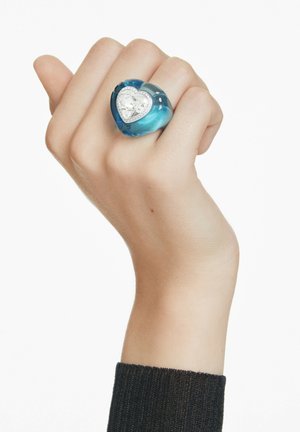 Una mano con una manga oscura lleva un anillo grueso azul que presenta una gran piedra preciosa transparente en forma de corazón y piedras más pequeñas que la rodean.