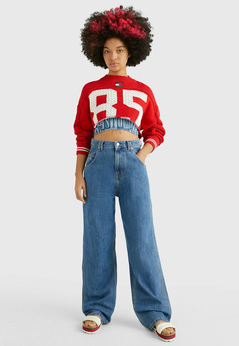 Maglione rosso lavorato a maglia con grande grafica bianca "85", abbinato a jeans a vita alta blu a gamba larga. Il modello indossa sandali bianchi con piattaforma.
