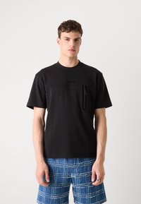Svart bomull t-skjorte med korte ermer, glidelåslomme på brystet og preget logo. Modellen har på seg blå mønstrede shorts.
