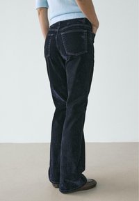 Jeans a zampa di colore blu scuro con finitura testurizzata, dotati di tasche posteriori e design a vita alta. Indossati con una maglia azzurro chiaro.