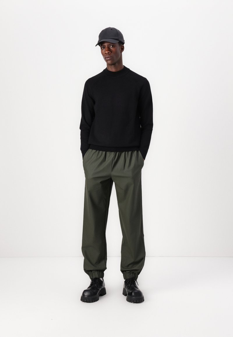 Schwarzer Strickpullover, olivgrüne Hose mit elastischem Bund und schwarze klobige Schnürstiefel; Model trägt eine graue Baseballmütze.