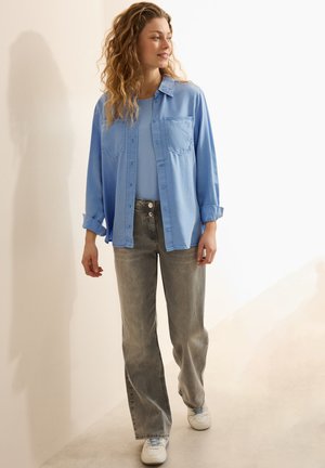Light blue button-up shirt with two chest pockets, paired with gray flared jeans and white sneakers. Soft texture, relaxed fit.