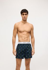 Pánské boxerky v tmavě zelené barvě s motivem malých bílých log. Mají bílý elastický pas s potiskem "LACOSTE" a zapínání na knoflík.