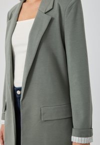 Blazer vert clair au design ajusté, avec une poche avant et des poignets retroussés révélant un motif rayé. Tissu doux, revers structurés.