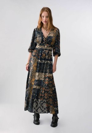 Femme aux longs cheveux portant une robe maxi patchwork florale et des bottines noires, debout devant un fond clair uni.