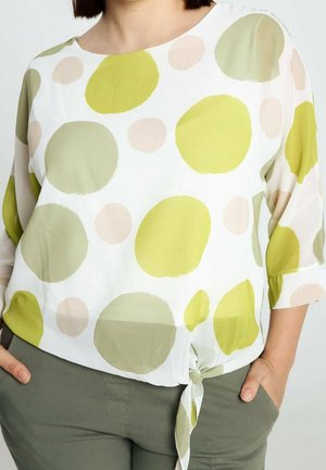 Kobieta w białej bluzce z dużymi zielonymi i beżowymi groszkami, zawiązanej w talii, w połączeniu z oliwkowozielonymi spodniami, z rękami w kieszeniach.