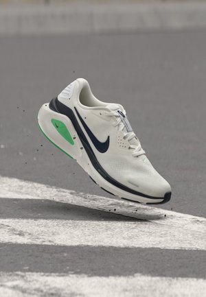 Λευκό αθλητικό παπούτσι Nike με μαύρο σήμα και πράσινες λεπτομέρειες, απαθανατισμένο στον αέρα πάνω από διάβαση πεζών σε γκρίζο πλακόστρωτο.
