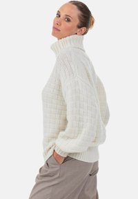 Grober, gestrickter Pullover in Creme mit hohem Kragen, der ein strukturiertes Muster und gerippte Bündchen aufweist. Kombiniert mit hellen Hosen.