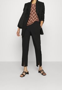 Musta räätälöity bleiseri, jonka päällä on sahalaitakuosiset ruskeat ja oranssit paidat, yhdistettynä mustiin lyhyisiin housuihin, joissa on sivuleikkaukset, ja mustiin nauhattomiin sandaaleihin.