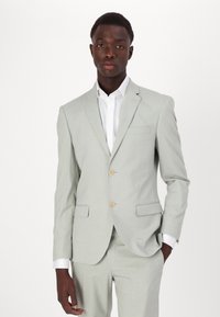 Šviesiai pilkas kostiumas su dviem mygtukais, liekna silueto išvaizda, įpjovos apykakle ir šoninėmis kišenėmis. Suderintas su balta marškiniais, lygaus tekstūros ir pritaikyto išvaizdos.