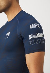 Tamsiai mėlyna sportinė marškinė su trumpomis rankovėmis, su UFC logotipu ir Venum prekės ženklu ant rankovės. Matomas tekstūruotas raštas medžiagoje.
