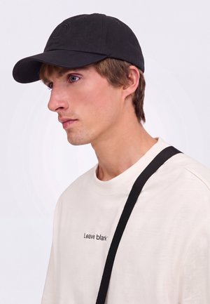 Jeune homme portant une casquette noire et un sweat-shirt blanc avec le texte "Leave blank", regardant vers la gauche sur un fond clair uni.