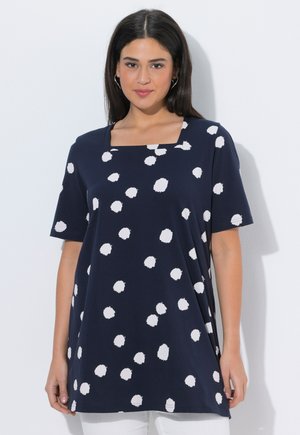 Kvinde med lyst, glat, mørkt hår iført marineblå tunika med uregelmæssige hvide prikker og firkantet udskæring, stående mod hvid baggrund.