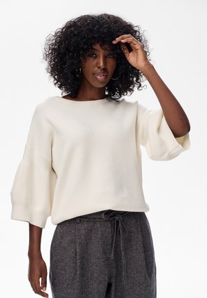 Crèmekleurige gebreide trui met brede, cropped mouwen. De stof lijkt zacht en textuur te hebben, gecombineerd met donkere, gestructureerde broeken.