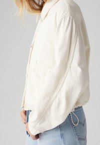 Vit canvasjacka med krage, sänkta axlar, sidfickor och en åtspänd fålldetalj. Bärs över ljusblå denim.