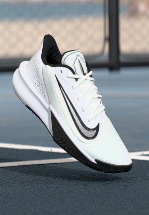 Hvide og sorte Nike løbesko med struktureret sål, der svæver over en blå sportsbane nær et kædhegn.