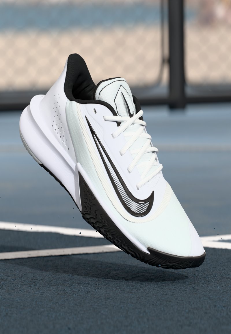 Hvide og sorte Nike løbesko med struktureret sål, der svæver over en bl�å sportsbane nær et kædhegn.