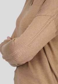 Pull à manches longues en tissu beige clair. Présente une texture douce avec des détails de couture visibles le long des manches. Encolure ronde.