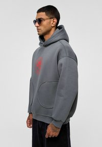 Grijze oversized hoodie met een rood grafisch ontwerp, grote voorzakken en een buidelzak, gemaakt van zachte gestructureerde stof.