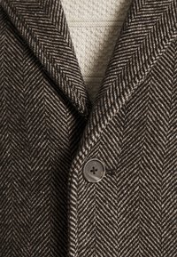 Primo piano di un cappotto a motivo zig-zag marrone e beige abbottonato su un maglione a maglia cremisi testurizzato.