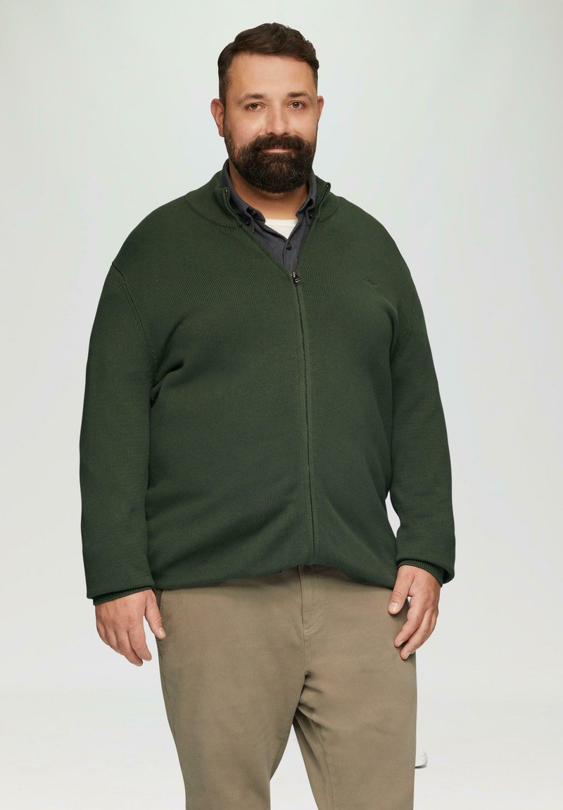 Dunkelgrüne Zip-Up-Strickjacke mit gerippter Textur, ausgestattet mit einem hohen Kragen und langen Ärmeln, kombiniert mit hellbeigen Hosen.