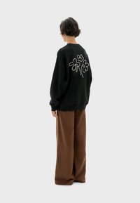 Zwarte oversized sweatshirt met witte bloemenborduurwerk op de achterkant, gecombineerd met losse bruine broeken van een zachte, textuurachtige stof.