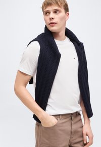 Νέος άνδρας με κοντά ξανθά μαλλιά, φορώντας λευκό μπλουζάκι με στρογγυλή λαιμόκοψη, μπεζ παντελόνι και μπλε σκούρα ζακέτα πλεκτή με σχέδιο καλώδιο περασμένη στους ώμους, κοιτάζοντας προς τα αριστερά.