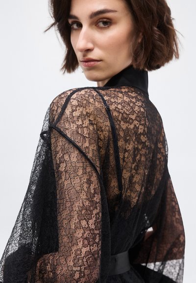 Mujer con cabello corto y castaño que lleva una blusa transparente de encaje floral negra con cuello alto, mirando por encima del hombro contra un fondo blanco.