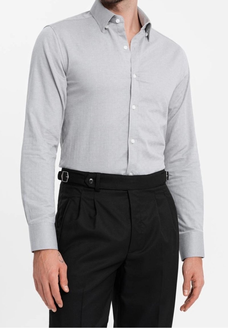 Homme portant une chemise gris clair à boutons avec manches longues et un pantalon noir taille haute avec des boucles de réglage latérales.