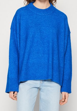 Personne portant un pull oversize bleu vif avec des poignets et un bas côtelés, associé à un jean bleu clair sur un fond uni.