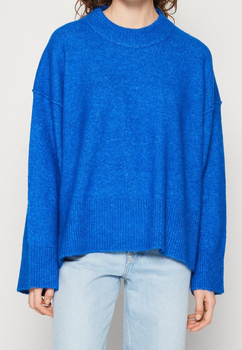 Personne portant un pull oversize bleu vif avec des poignets et un bas côtelés, associé à un jean bleu clair sur un fond uni.