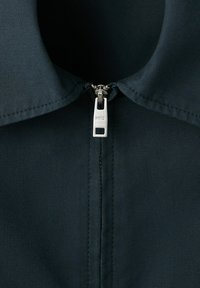 Close-up of a dark fabric collar with a silver zipper pull marked "MNG" centered at the top of a fully closed zipper.
