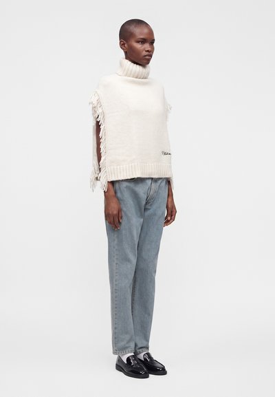 Femme portant un pull sans manches blanc à col roulé avec des franges sur les côtés, un jean bleu clair, des chaussettes blanches et des mocassins noirs, debout devant un fond uni.