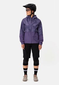 Asmuo, dėvintis purpurinę dviračių striukę, juodus šortus, kelių apsaugas, šalmą, akinius ir smėlinius batus, stovintis veidu šiek tiek į kairę.