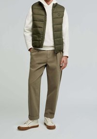 Olivgrüner Steppweste über einem weißen Hoodie, kombiniert mit beigen Hosen und cremfarbenen Sneakers. Minimalistisches Design mit seitlichen Taschen.