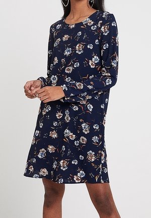 Kvinde iført en marinblå kjole med lange ærmer og et blomsterprint i hvide, brune og lyseblå nuancer.