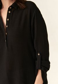 Black, textured button-up shirt with a collar, half-button front, and rolled sleeves, featuring wooden buttons and a loose fit.