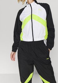 Svart och vit vindjacka med gröna accentlinjer, dragkedjestängning och elastiska ärmslut; tillsammans med matchande svarta byxor som har liknande designelement.
