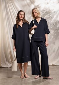 Deux mannequins portent des tenues modernes bleu marine foncé. L'un porte une robe ample jusqu'aux genoux, l'autre un haut avec un pantalon à jambes larges. Les deux présentent des coutures blanches.