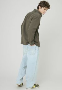 Chemise verte olive texturée avec un col, associée à un jean bleu clair ample et des baskets vertes avec des accents jaunes, vue en angle.