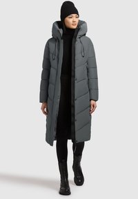 Lang, polstret grå jakke med hætte, der har en frontlynlås, trykknapper, sidelommer og en struktureret overflade. Models sorte tøj og støvler.