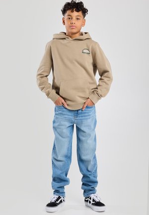 Sudadera beige con bolsillo frontal, con texto impreso en verde; combinada con jeans baggy de color azul claro y zapatillas negras.