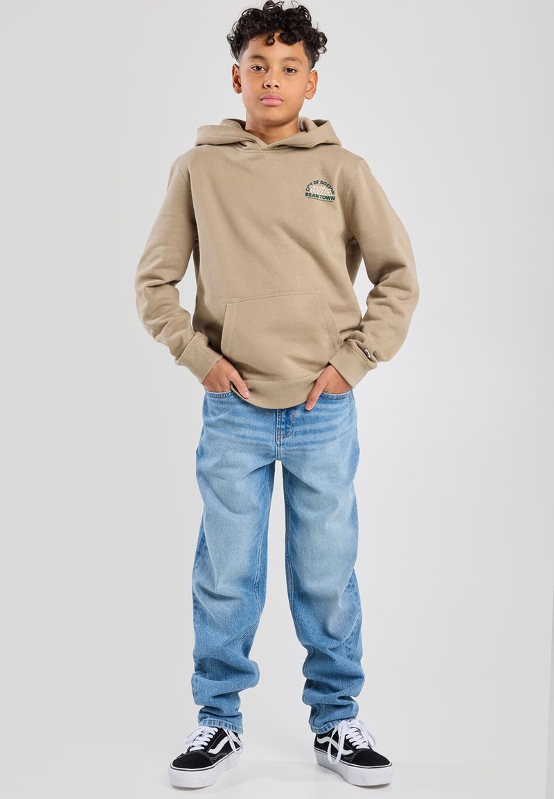 Sudadera beige con bolsillo frontal, con texto impreso en verde; combinada con jeans baggy de color azul claro y zapatillas negras.