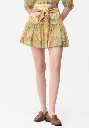 Frau trägt einen kurzen, plissierten Rock mit retro Blumenmuster und einem gebundenen Gürtel, kombiniert mit braunen Leder-Loafers vor einfachem Hintergrund.