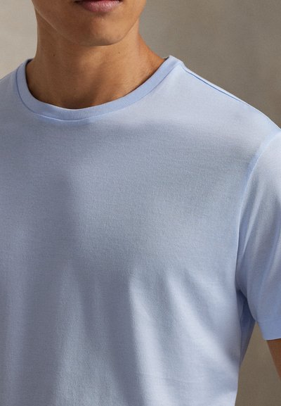 Κοντινό πλάνο ενός ατόμου που φοράει ανοιχτό μπλε t-shirt με στρογγυλή λαιμόκοψη και κοντά μανίκια, πάνω σε ουδέτερο φόντο.