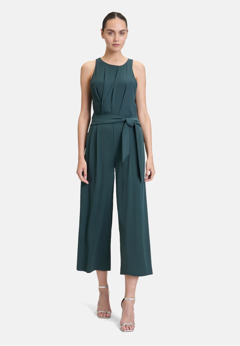 Tuta turchese con top aderente, pieghe e cintura legata. Presenta pantaloni larghi e a gamba corta e un tessuto liscio e testurizzato. Spalline trasparenti.