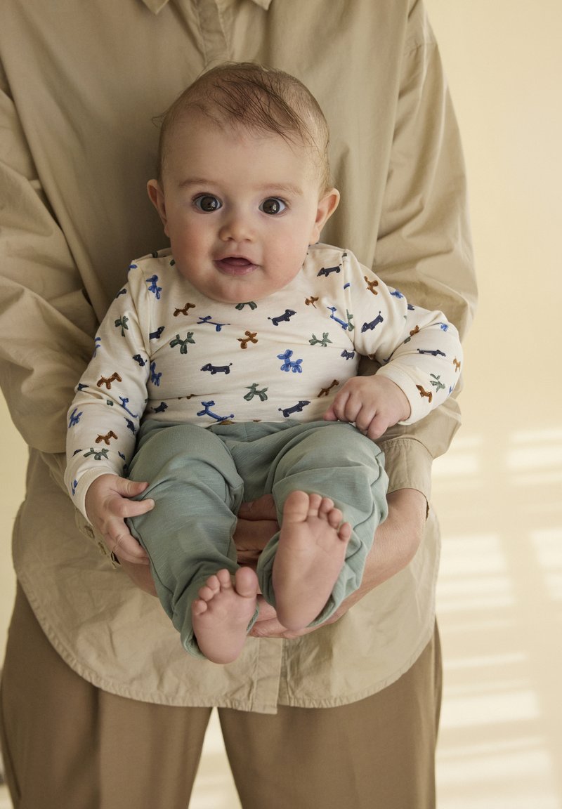 Bebé viste una camiseta de manga larga color crema con estampados de perros de colores y pantalones verdes suaves. Sostenido en brazos, con los pies descalzos visibles.